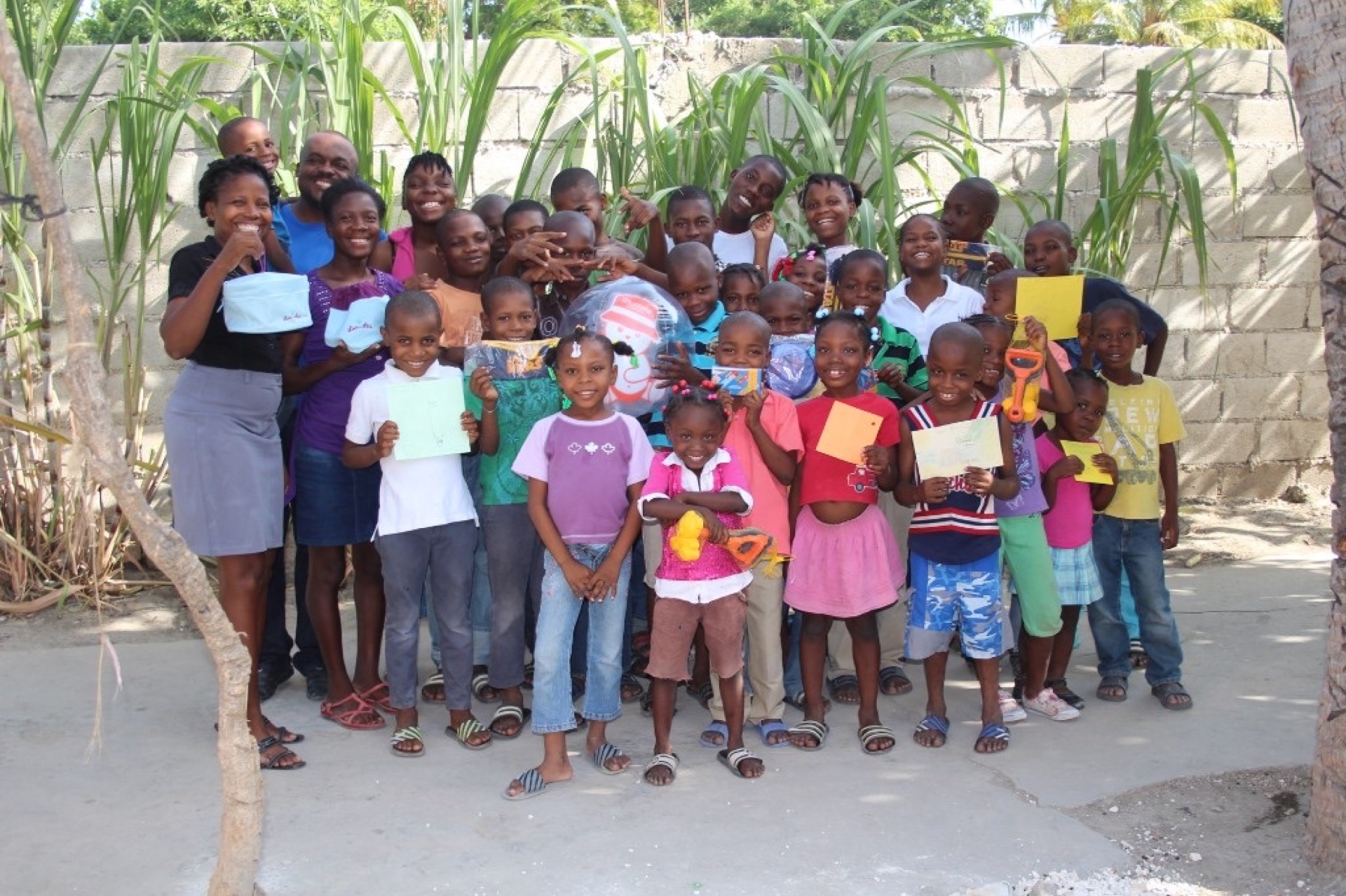 Groupe d'enfants et d'adultes en extérieur, souriant avec cartes et jouets devant un mur en parpaings